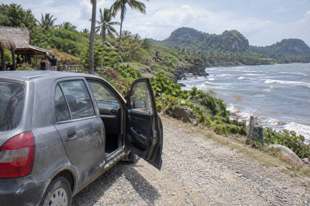 A parked rental car on a rural road with palm trees and green hills behind it