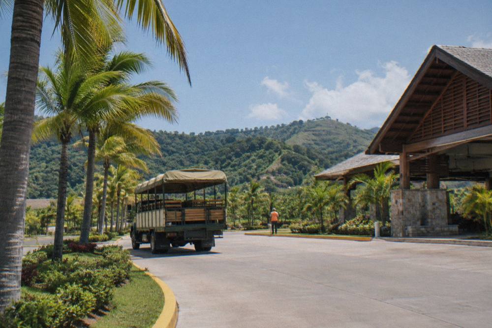 Green safari truck at a tarmac entrance with palms, a worker in orange vest, cruise terminal behind.