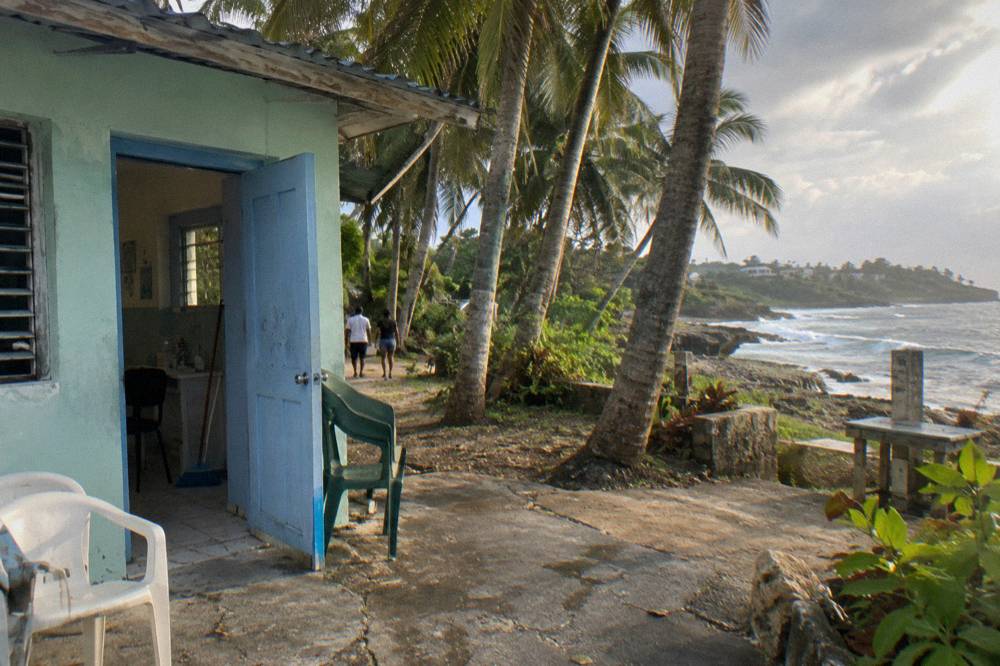 A simple rural clinic building with an open door and chairs outside under palm trees