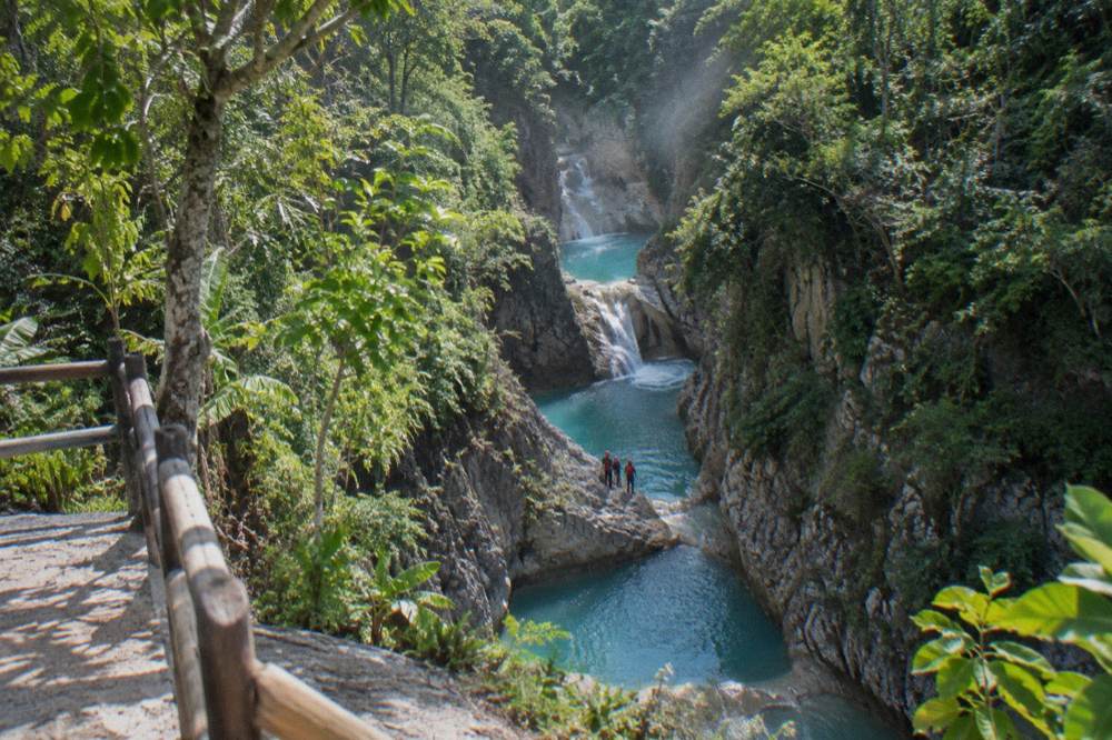 Turquoise cascade pools in a rocky jungle canyon, tiny figures in red helmets at pool edge, mist rising