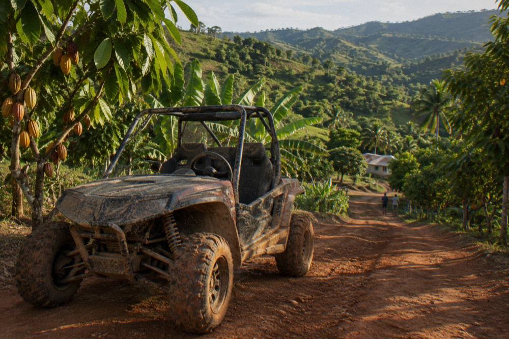 A mud caked two seat off road buggy parked on a red clay track between tropical trees and cacao