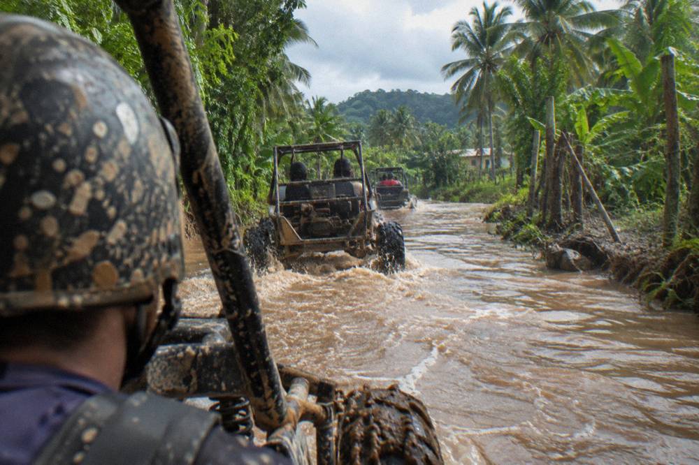 A line of off-road buggies driving along a muddy track through dense green palm forest