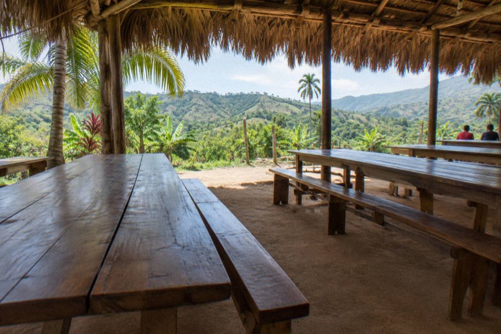 Open air thatched shelter with plank tables on packed earth, palm fronds shading, green hills beyond