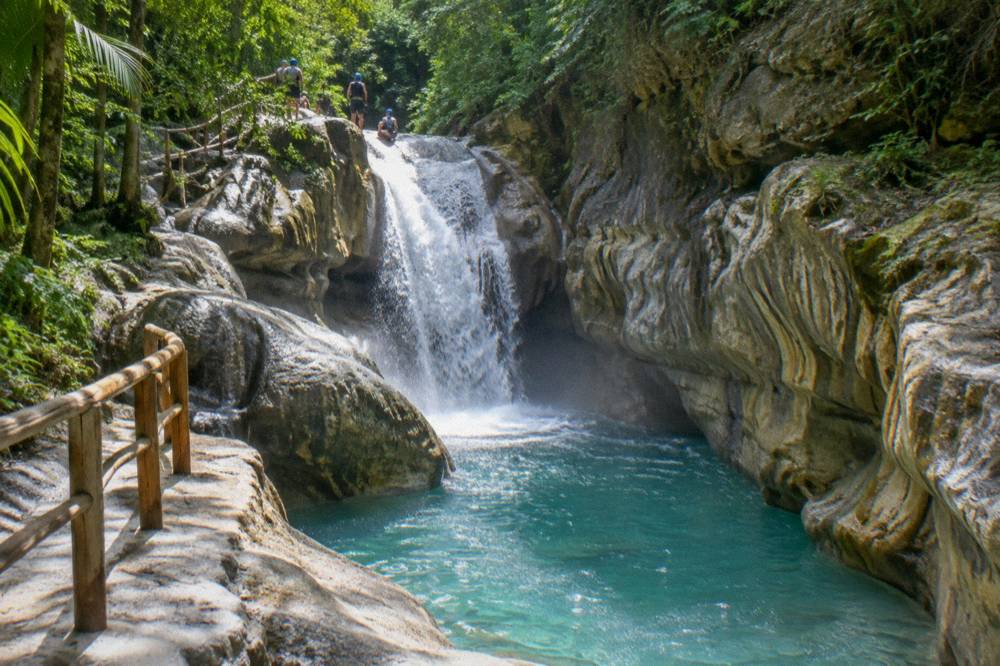 Waterfall pouring into a turquoise pool between smooth rock walls with tiny helmeted figures above