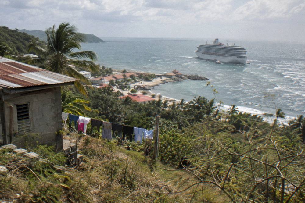 A cruise ship at anchor seen from a green hillside above a Caribbean bay