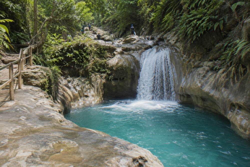 Turquoise pool under a pouring cascade in a limestone canyon, wet rock shelves, helmeted figures above.